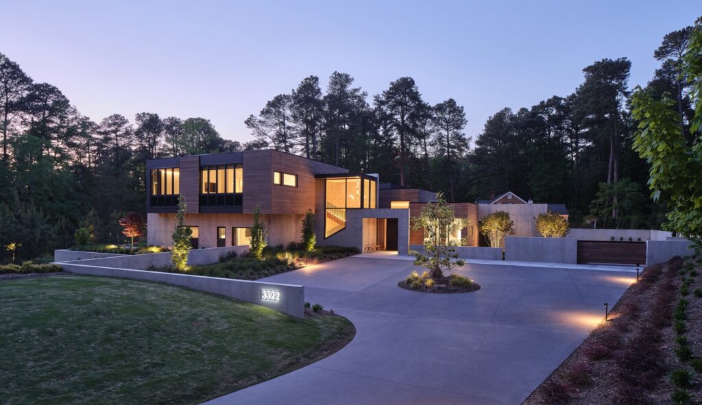 Modern Durham NC home at dusk with warm lit windows, cedar and concrete facade and a curved concrete motor court